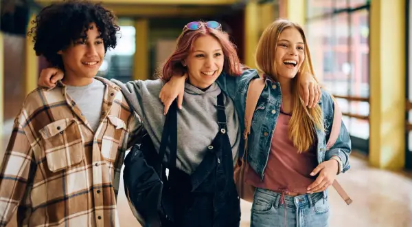 Drei lachende Freunde gehen Arm in Arm durch einen Flur. Sie sind im perfekten Alter, um zu sagen: ab wann ist man jugendlich?