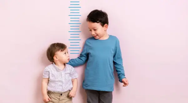 Zwei Jungen stehen vor einer Messlatte. Der ältere Junge, vielleicht 4 Jahre alt, legt dem kleineren seine Hand auf die Schulter.