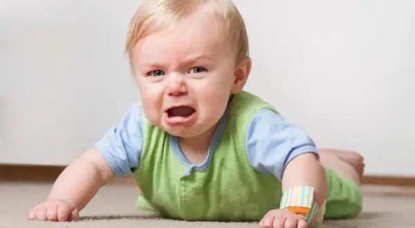 Ein weinender **baby 16 wochen** alt liegt auf dem Bauch auf einem Teppich. Es hat blonde Haare und trägt ein grünes Oberteil über einem blauen Hemd.