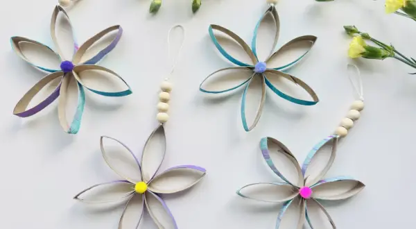 Bunte Blumen aus Klopapierrollen basteln: Vier hübsche Anhänger mit Holzperlen und Pompons, umgeben von gelben Blüten.