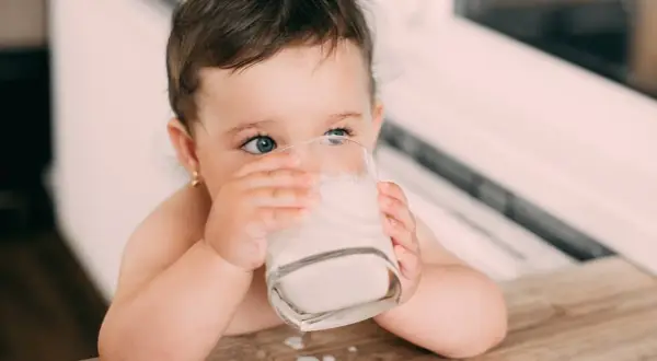 Kleines Kind trinkt aus einem Glas Milch. Die blauen Augen blicken neugierig. Baby Kuhmilch ist wichtig für das Wachstum.