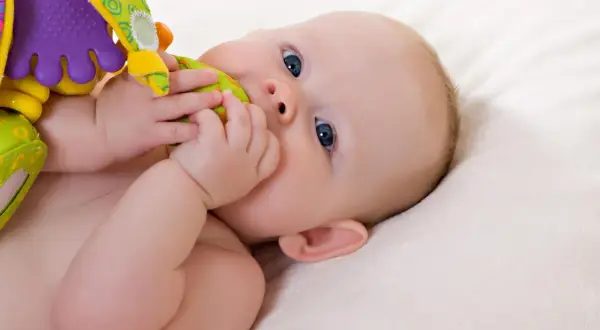 Ein **baby 19 wochen** alt mit blauen Augen liegt auf einer weichen Decke und kaut auf einem bunten Spielzeug.