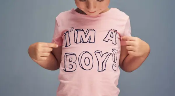 Ein Kind mit Mütze zeigt stolz auf sein rosa T-Shirt mit der Aufschrift "I'M A BOY!". Ein Bild, das die Identität von transgender Kindern feiert.