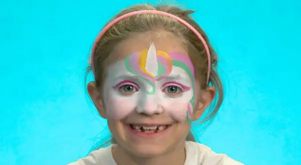 Ein Mädchen mit einem bunten Einhorn-Schminken für Kinder. Das Gesicht ist weiß grundiert, mit einem weißen Horn und bunten Bögen in Rosa, Gelb und Grün.
