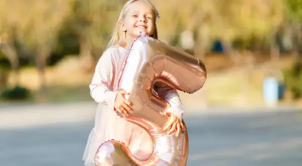 Ein glückliches, blondes Kind von 5 Jahren hält stolz einen großen, roségoldenen Ballon in Form der Zahl 5.