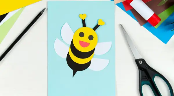 Süße Biene basteln: Gelber Kopf mit Herzfühler, schwarze Streifen, weiße Flügel auf hellblauem Papier. Schere und Klebestift liegen bereit.
