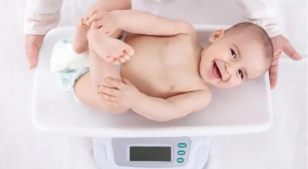 Ein lächelndes Baby liegt auf einer Waage für die U3 Untersuchung. Hände halten das Kind sanft.