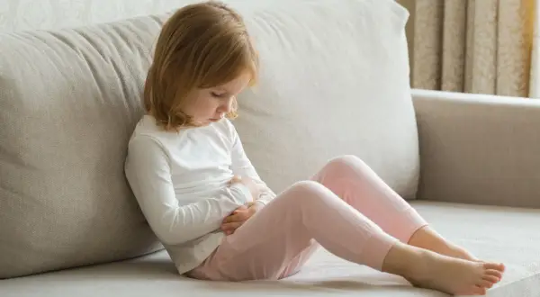 Ein Kleinkind mit blonden Haaren sitzt auf einer Couch und hält sich den Bauch. Es scheint, als hätte das Kleinkind Erbrechen ohne weitere Symptome.