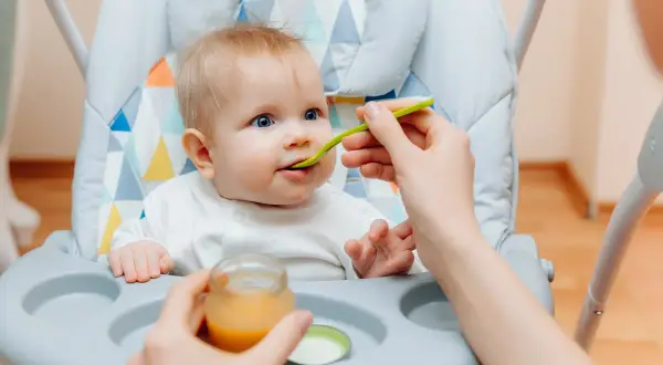 Ein süßes Baby von 6 Monaten sitzt im Hochstuhl und bekommt Brei gefüttert.