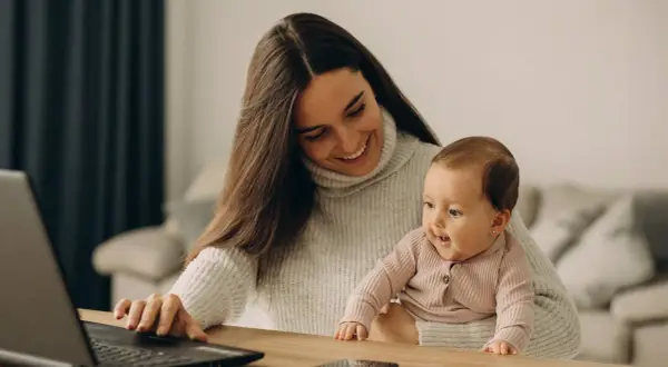 Lächelnde Mutter mit Baby auf dem Schoß am Laptop. Ein Bild, das den Spagat zwischen Beruf und Familie zeigt, oft verbunden mit dem Gefühl von Mama Burnout.