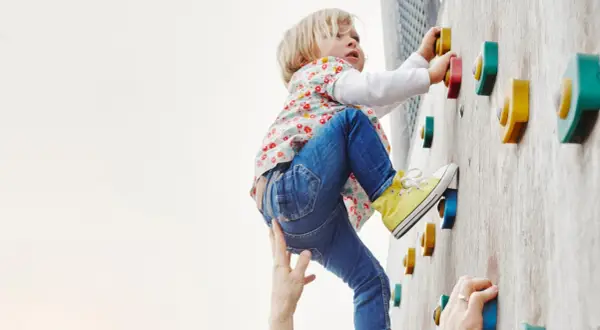 Kleines Kind klettert an einer Boulderwand, fördert seine Kompetenz. Hände helfen und sichern.