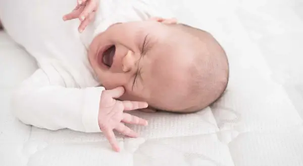 Ein Baby mit weit geöffnetem Mund und geschlossenen Augen, das sich im unruhigen Schlaf windet. Seine kleinen Hände sind nach oben gestreckt.