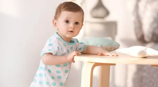 Ein neugieriges Baby, etwa 1 Jahr alt, stützt sich auf einen Holztisch. Es trägt ein weißes T-Shirt mit blauen Tupfen.