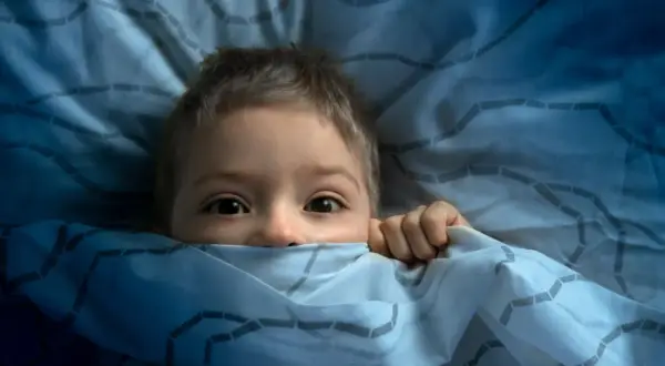 Ein Kind versteckt sich ängstlich unter der Decke. Ab wann haben Kinder Angst im Dunkeln?