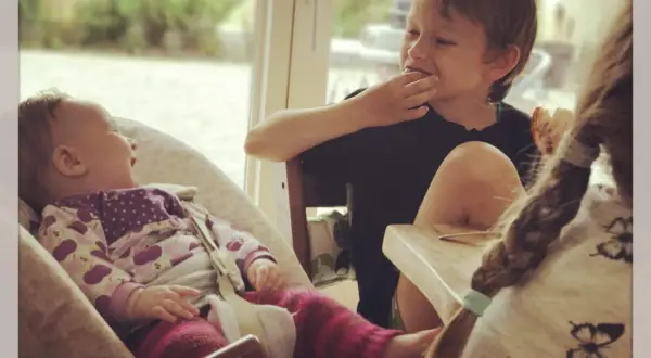 Drei Kinder am Tisch: Ein Baby lacht, ein Junge isst, ein drittes Kind mit Zopf ist im Vordergrund.