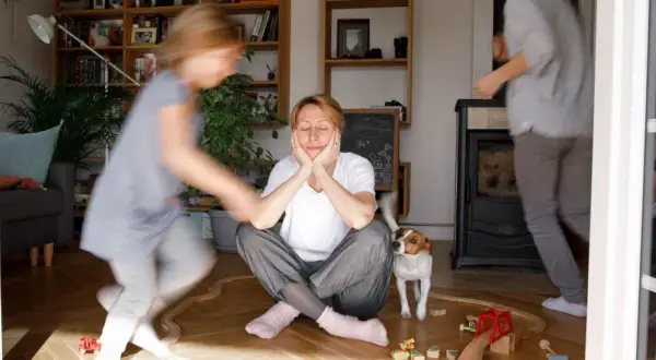 Chaos im Wohnzimmer: Kinder rennen herum, ein Hund schaut neugierig, und die mama steht kopf. Sie sitzt auf dem Boden, die Augen geschlossen, während das Leben um sie herum tobt.