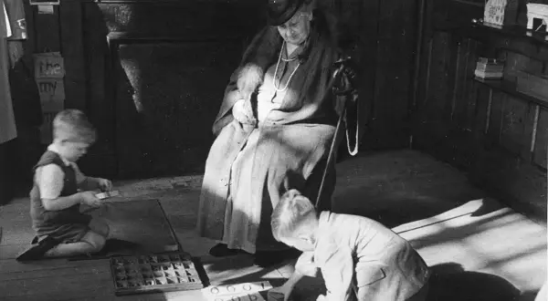 Maria Montessori beobachtet Kinder beim Lernen. Ein Junge kniet und sortiert Buchstaben, ein anderer übt mit Zahlen.