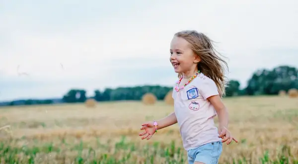 Ein fröhliches Kleinkind rennt lachend über ein Feld. Seine blonden Haare wehen im Wind, und es trägt ein buntes Halsband.