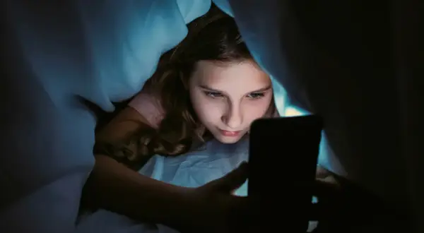 Ein Mädchen liegt im Bett unter der Decke und schaut auf ihr Handy. Sie wirkt vertieft, fast wie faule Kinder, die ihre Zeit mit digitalen Medien verbringen.