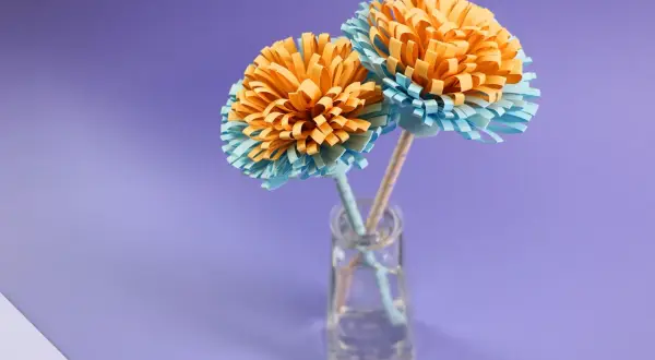 Zwei selbstgebastelte Papierblumen in Orange und Hellblau stehen in einer kleinen Glasvase. Ein schönes Beispiel für blumenstrauß basteln.