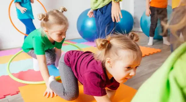 Kinder haben Spaß bei Bewegungsspielen im Kindergarten. Sie krabbeln auf bunten Matten, umgeben von Gymnastikbällen und Reifen.