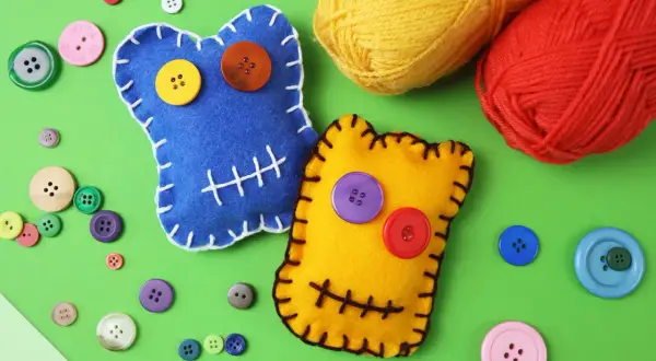 Zwei bunte Filz-Wutmonster basteln: Ein blaues mit gelbem und orangefarbenem Knopfauge, ein gelbes mit lila und rotem Knopfauge. Viele bunte Knöpfe und Garnknäuel liegen daneben.