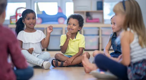 Kinder sitzen im Kreis und lernen. Die Lehrerin beobachtet aufmerksam, um die **Schulreife erkennen** zu können.