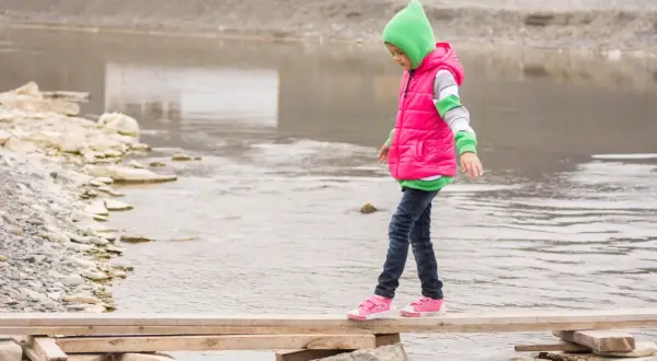 Ein Kind balanciert über ein provisorisches Holzbrett über einem Fluss. Das zeigt, was bedeutet selbstwirksamkeit: Mut, Vertrauen in die eigenen Fähigkeiten und das Überwinden von Hindernissen.