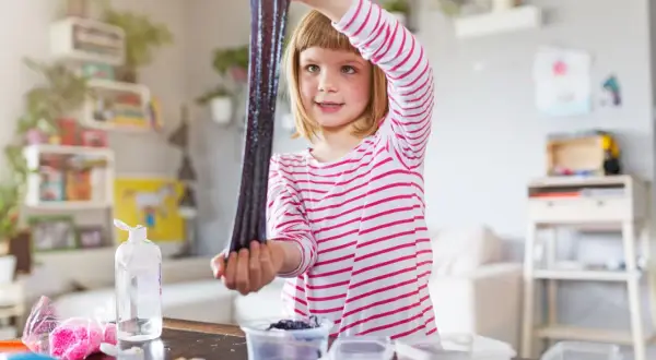 Ein Mädchen hält glitzernden, dunklen Schleim hoch, den sie gerade selber gemacht hat. Auf dem Tisch liegen Zutaten für weitere Schleim-Kreationen.