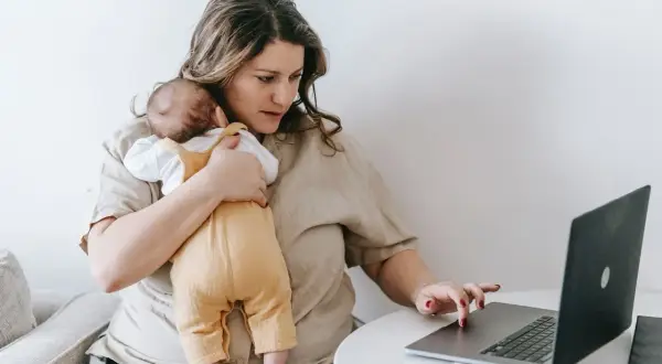 Alleinerziehend und im **burnout**: Eine Frau hält ihr Baby und arbeitet am Laptop.