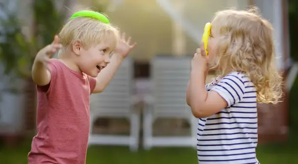 Zwei Kinder spielen im Garten. Der Junge mit einem grünen Ring auf dem Kopf lacht, das Mädchen hält ein gelbes Spielzeug. Ein Beispiel für gelebte sozialkompetenz definition.