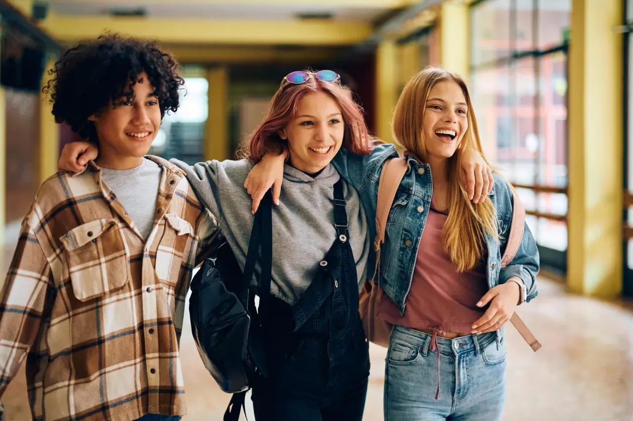 Drei lachende Freunde gehen Arm in Arm durch einen Flur. Sie sind im perfekten Alter, um zu sagen: ab wann ist man jugendlich?