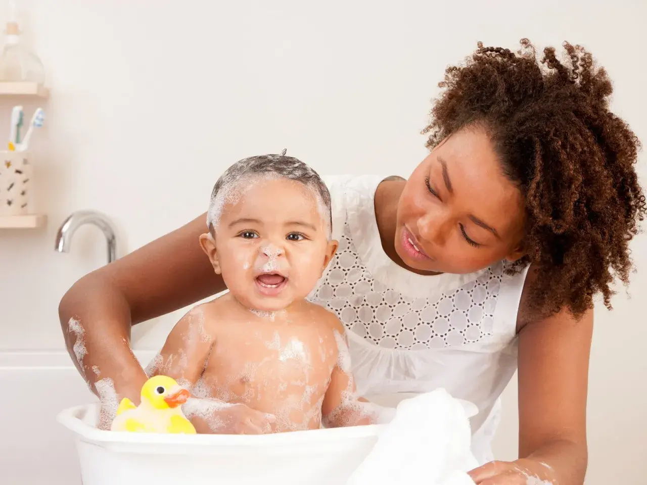 Mutter wäscht ihr Baby mit Schaum und Quietscheente. Ein warmes Bad kann bei Fieber helfen.