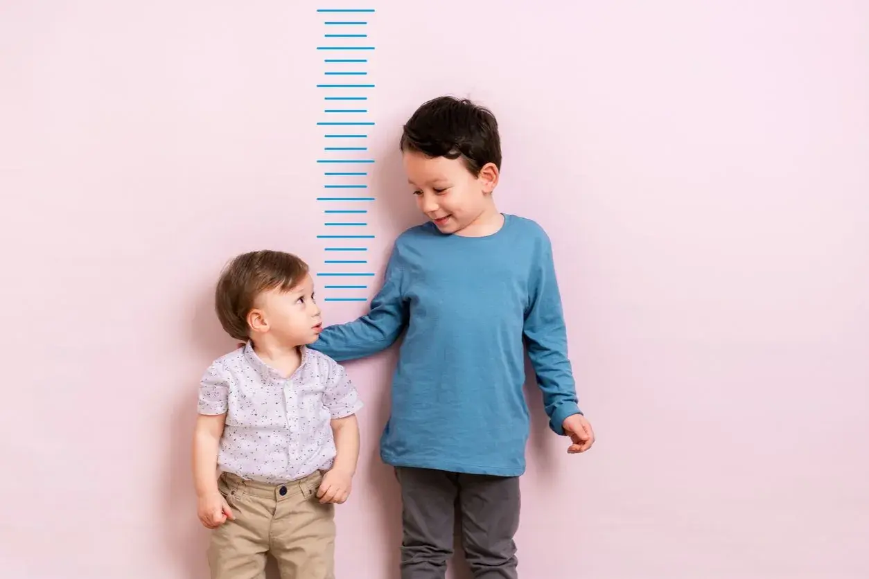 Zwei Jungen stehen vor einer Messlatte. Der ältere Junge, vielleicht 4 Jahre alt, legt dem kleineren seine Hand auf die Schulter.