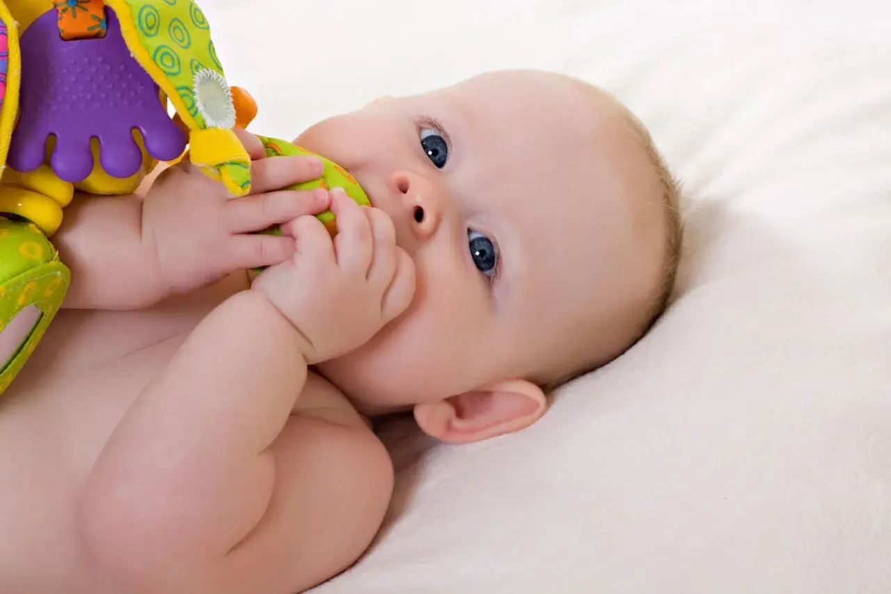 Ein **baby 19 wochen** alt mit blauen Augen liegt auf einer weichen Decke und kaut auf einem bunten Spielzeug.