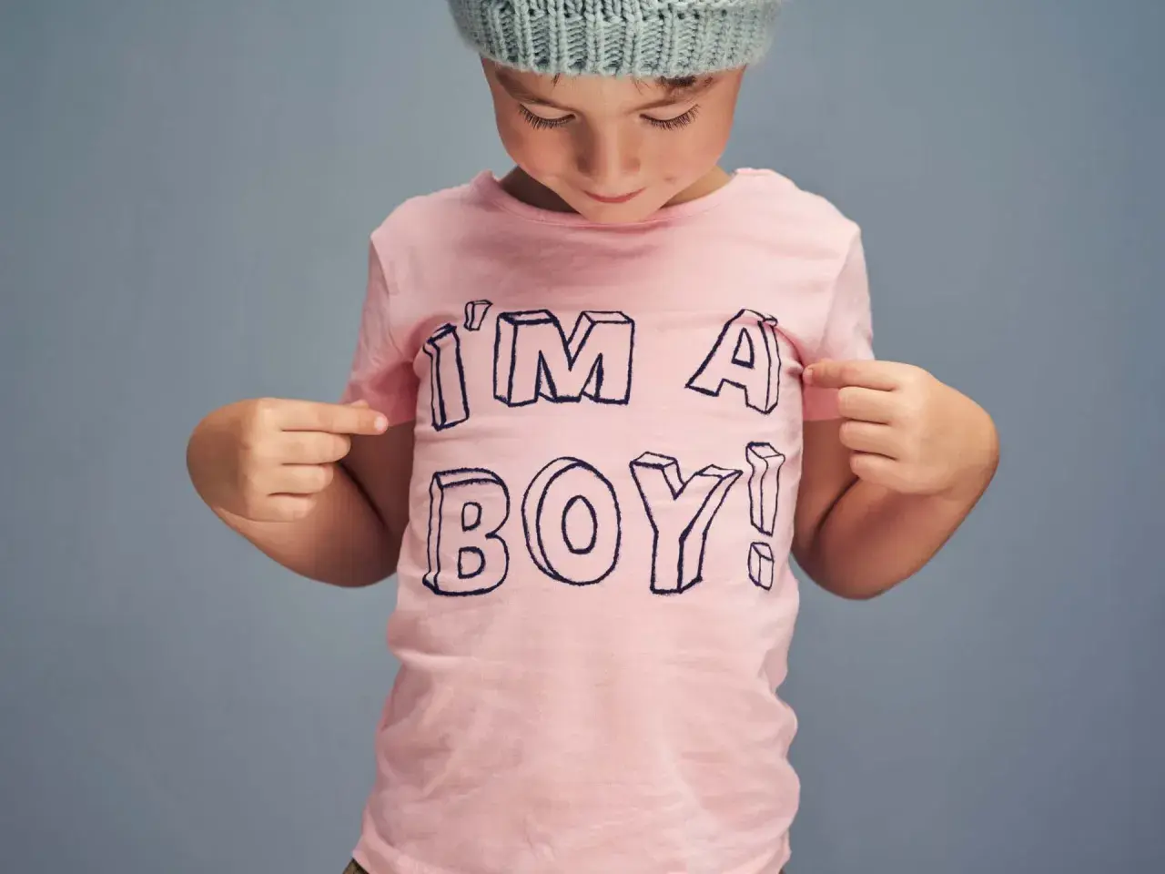 Ein Kind mit Mütze zeigt stolz auf sein rosa T-Shirt mit der Aufschrift "I'M A BOY!". Ein Bild, das die Identität von transgender Kindern feiert.