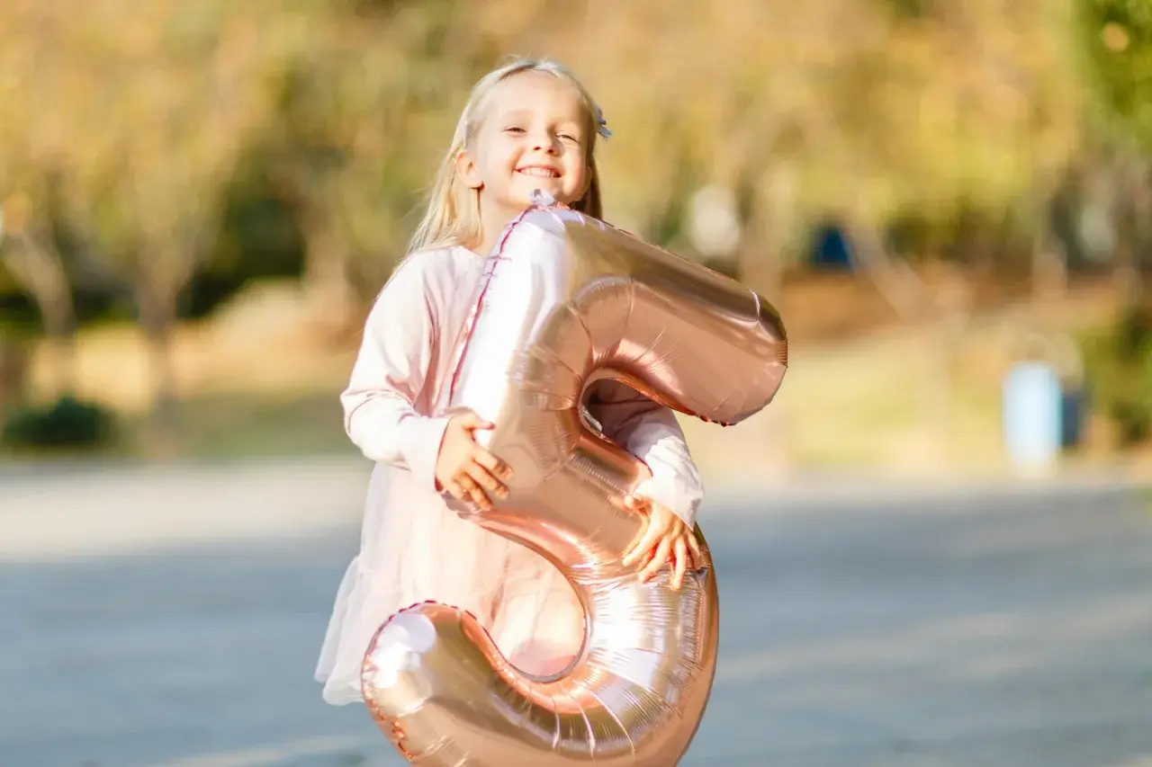 Ein glückliches, blondes Kind von 5 Jahren hält stolz einen großen, roségoldenen Ballon in Form der Zahl 5.