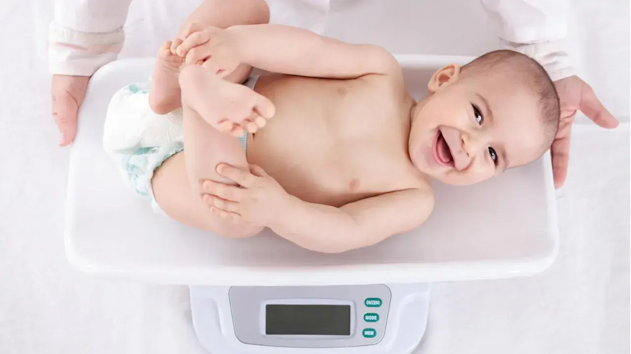 Ein lächelndes Baby liegt auf einer Waage für die U3 Untersuchung. Hände halten das Kind sanft.