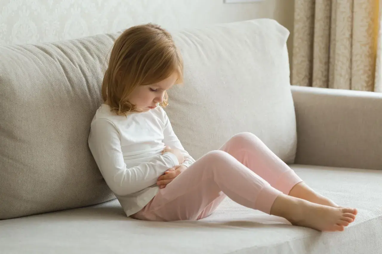 Ein Kleinkind mit blonden Haaren sitzt auf einer Couch und hält sich den Bauch. Es scheint, als hätte das Kleinkind Erbrechen ohne weitere Symptome.