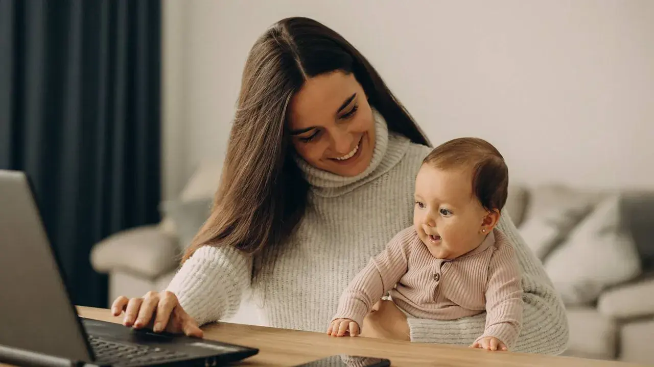 Lächelnde Mutter mit Baby auf dem Schoß am Laptop. Ein Bild, das den Spagat zwischen Beruf und Familie zeigt, oft verbunden mit dem Gefühl von Mama Burnout.