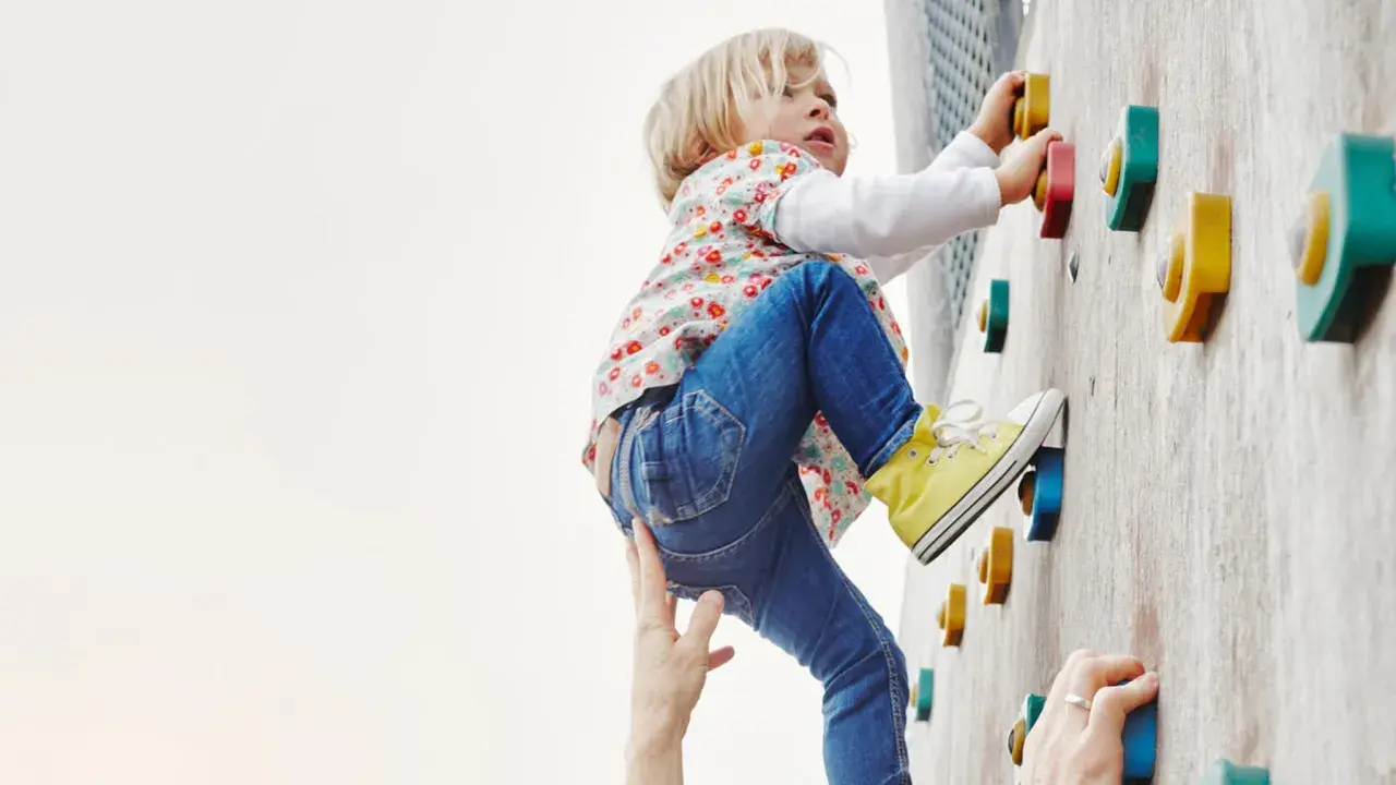Kleines Kind klettert an einer Boulderwand, fördert seine Kompetenz. Hände helfen und sichern.