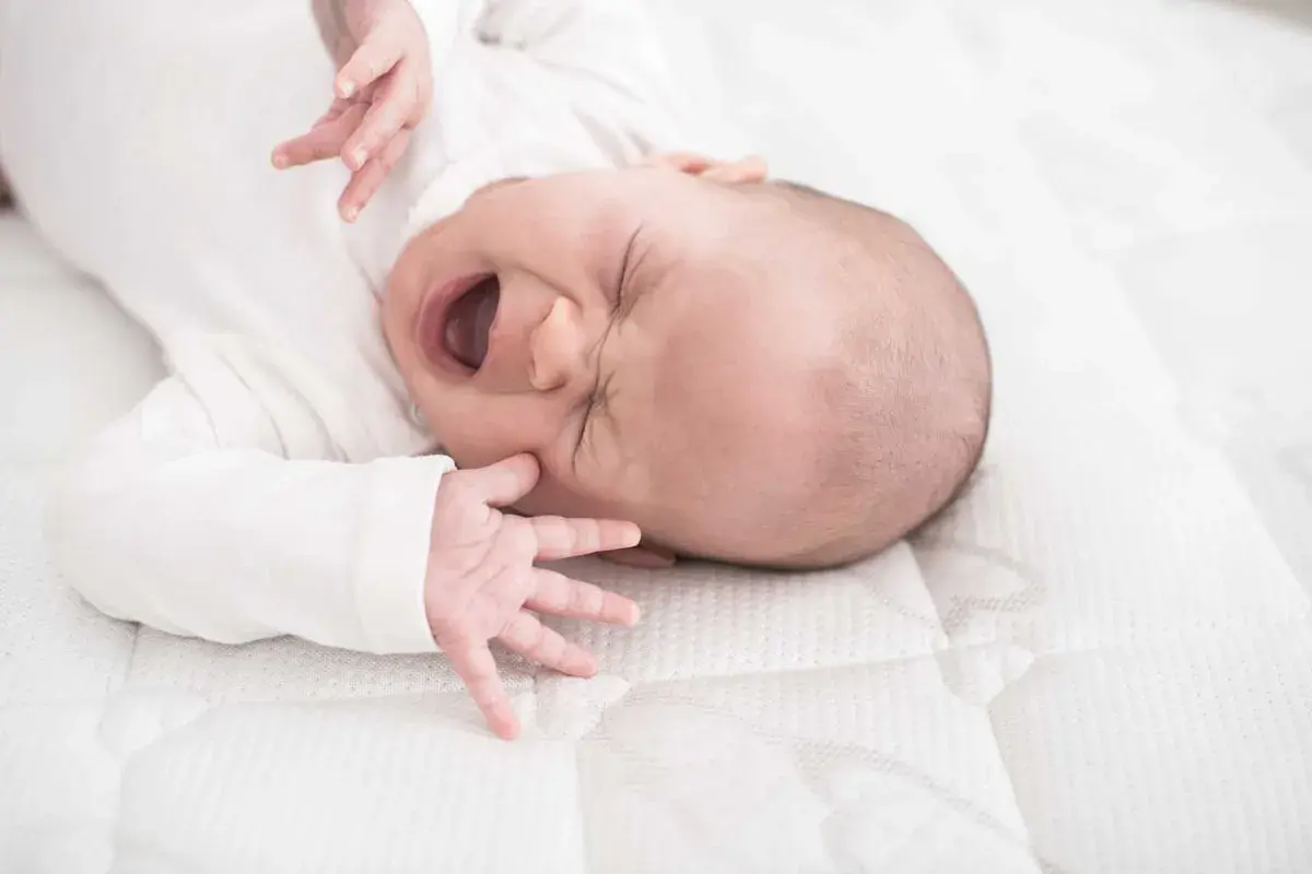 Ein Baby mit weit geöffnetem Mund und geschlossenen Augen, das sich im unruhigen Schlaf windet. Seine kleinen Hände sind nach oben gestreckt.