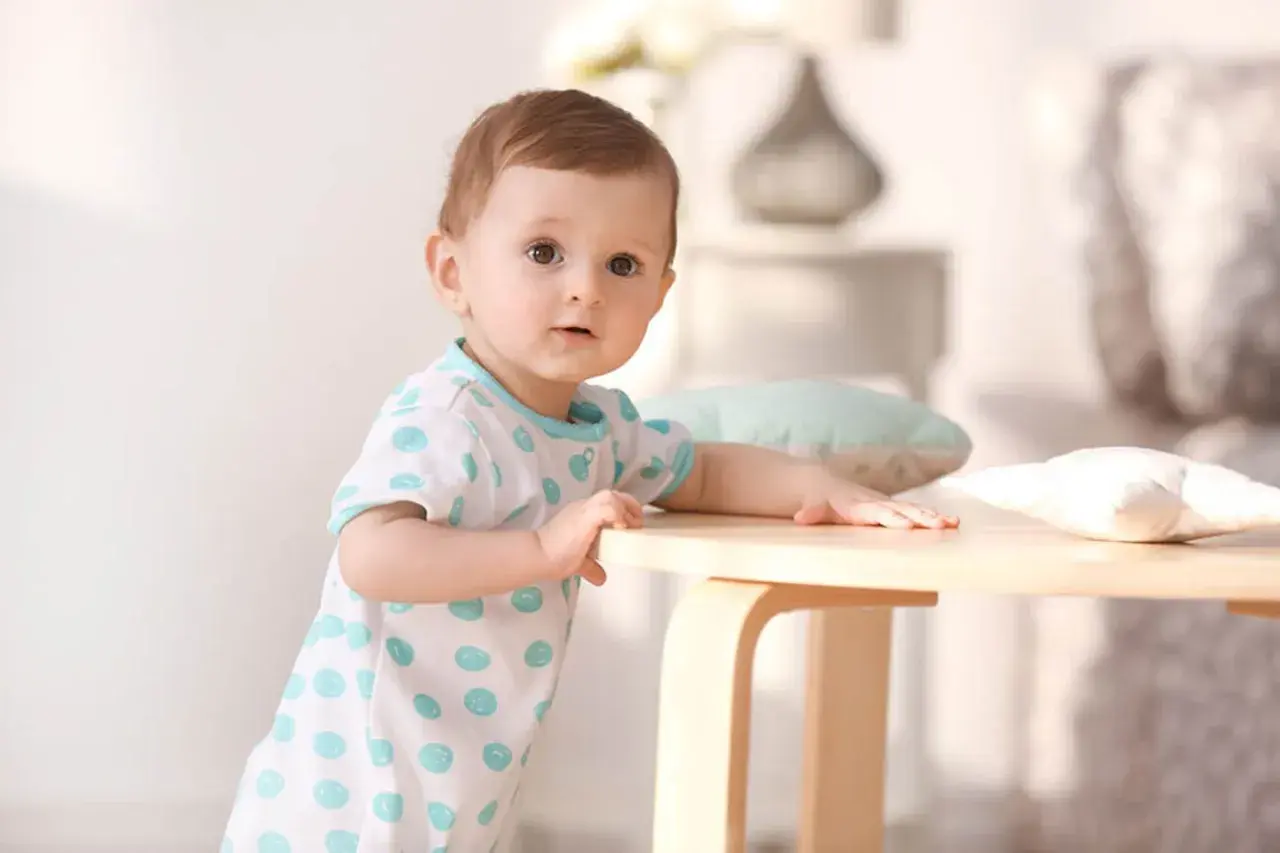 Ein neugieriges Baby, etwa 1 Jahr alt, stützt sich auf einen Holztisch. Es trägt ein weißes T-Shirt mit blauen Tupfen.