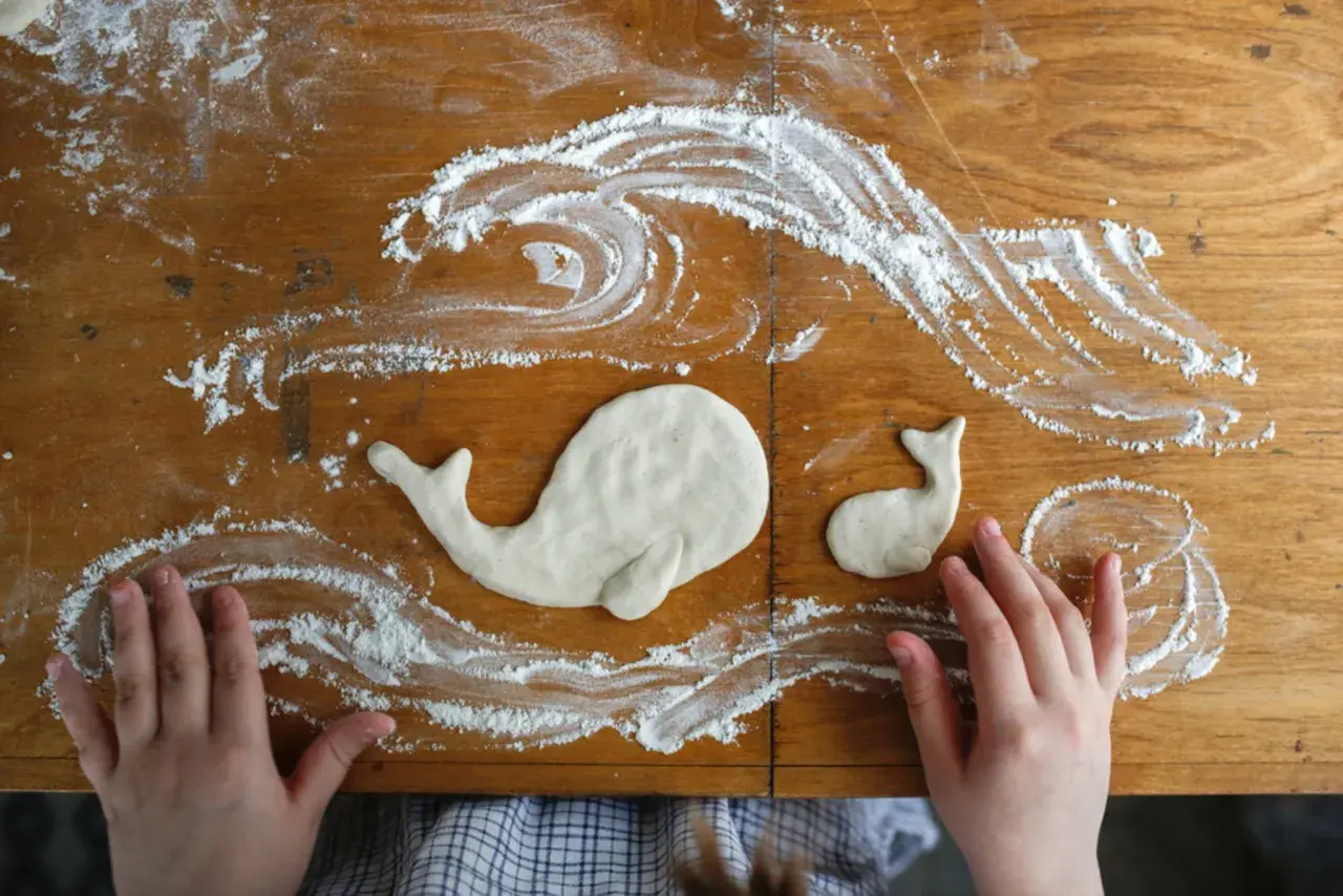 Kinderhände gestalten Salzteig-Ideen: zwei Wale schwimmen in Meereswellen aus Mehl auf einem Holztisch.