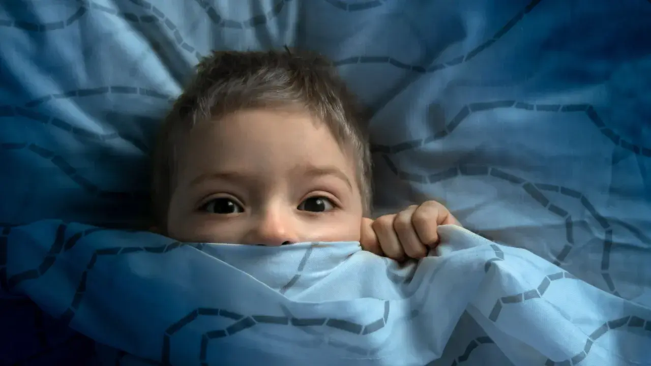 Ein Kind versteckt sich ängstlich unter der Decke. Ab wann haben Kinder Angst im Dunkeln?