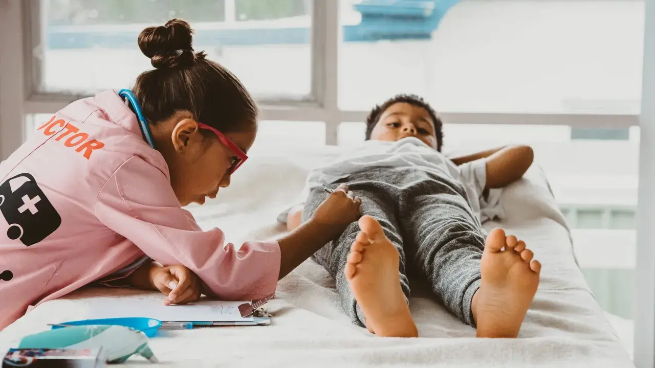 Kinder spielen Doktorspiele im Kindergarten. Ein Mädchen in Arztkittel untersucht einen Jungen, der auf einer Liege liegt.