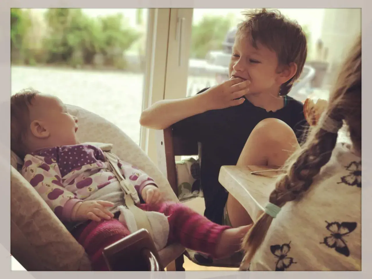 Drei Kinder am Tisch: Ein Baby lacht, ein Junge isst, ein drittes Kind mit Zopf ist im Vordergrund.