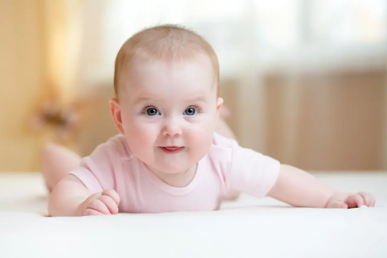 Ein süßes Baby mit blauen Augen liegt auf dem Bauch und versucht, den Kopf zu halten. Es trägt ein rosa Body.