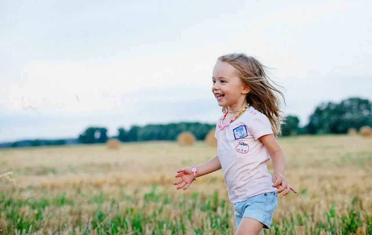 Ein fröhliches Kleinkind rennt lachend über ein Feld. Seine blonden Haare wehen im Wind, und es trägt ein buntes Halsband.