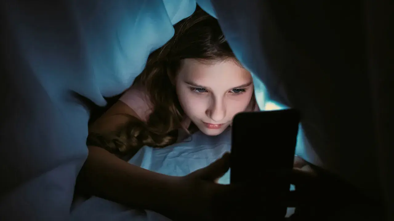 Ein Mädchen liegt im Bett unter der Decke und schaut auf ihr Handy. Sie wirkt vertieft, fast wie faule Kinder, die ihre Zeit mit digitalen Medien verbringen.